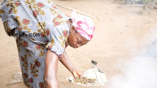 A woman in a wax print blouse and a red headtie stoops to feed palm kernels to a smoky fire. Image by Francesco DeAugustinis for Mongabay.